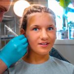 Girl in a dental chair receives a local anesthesia shot from a dentist wearing blue gloves; a friend or assistant is in the background.