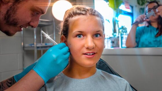 Girl in a dental chair receives a local anesthesia shot from a dentist wearing blue gloves; a friend or assistant is in the background.