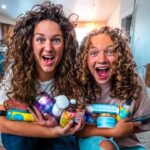 Two cheerful girls with curly hair hug a bunch of hair care products in a cozy living room.