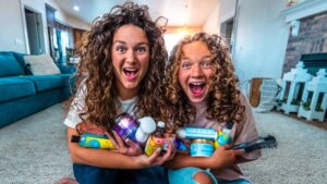 Two cheerful girls with curly hair hug a bunch of hair care products in a cozy living room.