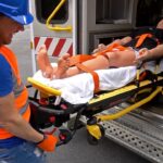 Emergency responder in an orange safety vest and blue helmet wheels a patient on a stretcher toward an ambulance entrance.