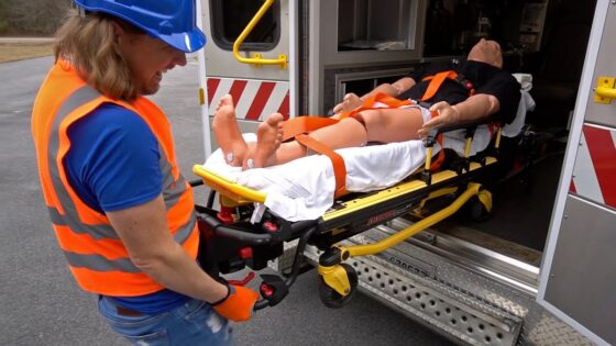Emergency responder in an orange safety vest and blue helmet wheels a patient on a stretcher toward an ambulance entrance.