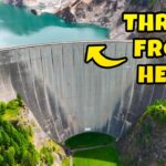 A massive curved concrete dam with turquoise water behind it and a large yellow sign reading 'THROW FROM HERE' on the upper right side.