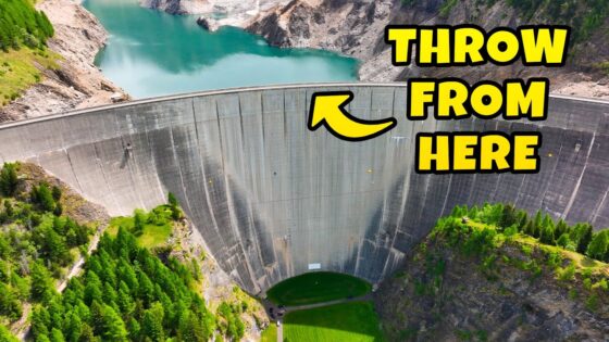 A massive curved concrete dam with turquoise water behind it and a large yellow sign reading 'THROW FROM HERE' on the upper right side.