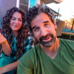 A man in a green shirt leans toward the camera while a smiling woman with curly hair holds a cracker beside him in a backyard setting with a trampoline in the background.