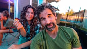 A man in a green shirt leans toward the camera while a smiling woman with curly hair holds a cracker beside him in a backyard setting with a trampoline in the background.