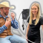 Man in a cowboy hat and brown leather vest sits on an exam chair, talking on a corded phone, with a smiling nurse in scrubs beside him in a clinic.