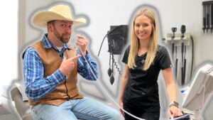 Man in a cowboy hat and brown leather vest sits on an exam chair, talking on a corded phone, with a smiling nurse in scrubs beside him in a clinic.