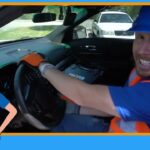 Driver wearing blue hard hat and orange safety vest, smiling, seated at the steering wheel inside a car; cartoon police frame and badge surround the image