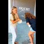Two girls barefoot on a porch, one shielding her face as the other playfully hits her with a tortilla; the word 'OUCH!' appears above.