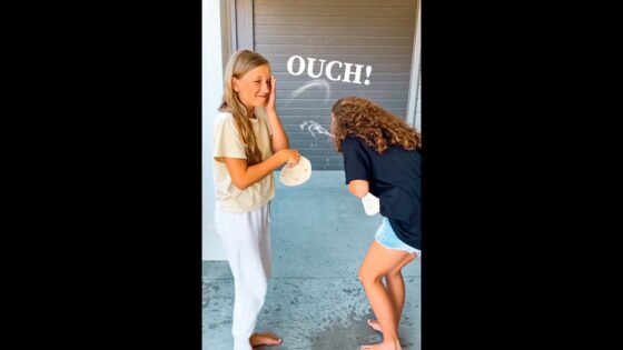 Two girls barefoot on a porch, one shielding her face as the other playfully hits her with a tortilla; the word 'OUCH!' appears above.