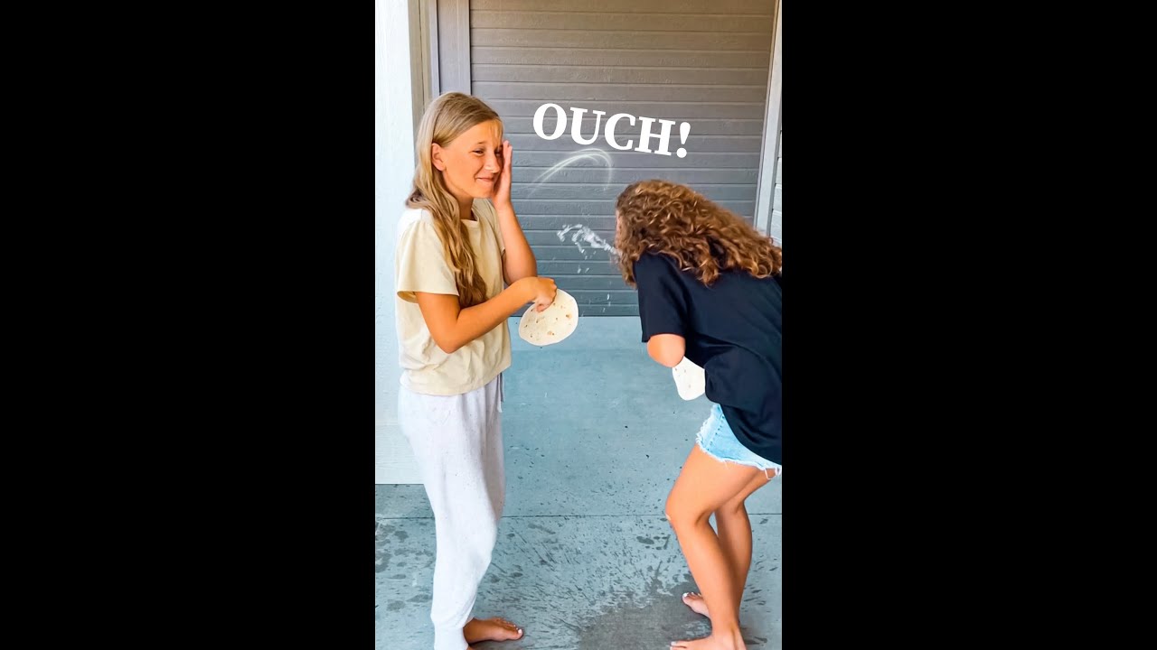 Two girls barefoot on a porch, one shielding her face as the other playfully hits her with a tortilla; the word 'OUCH!' appears above.