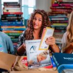 Three smiling girls sit among tall stacks of books, reading and sharing materials.