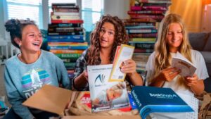 Three smiling girls sit among tall stacks of books, reading and sharing materials.