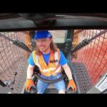 Construction worker in a blue helmet and orange safety vest sits in the cab of a heavy machine, hands on the controls, smiling at the camera.