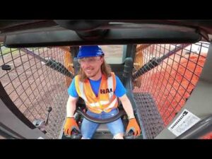 Construction worker in a blue helmet and orange safety vest sits in the cab of a heavy machine, hands on the controls, smiling at the camera.