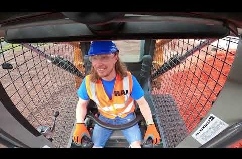Construction worker in a blue helmet and orange safety vest sits in the cab of a heavy machine, hands on the controls, smiling at the camera.