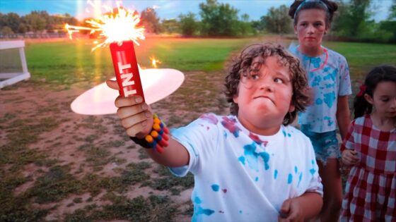 Young boy in a paint-splattered shirt holds a lit red TNT sparkler, sparks spraying as others watch in a park.