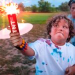 Child in a paint-splattered shirt holds a lit sparkler outdoors while friends watch.