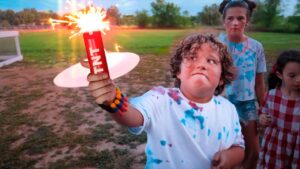 Child in a paint-splattered shirt holds a lit sparkler outdoors while friends watch.
