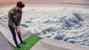 Man in a casual sweatshirt and dark pants stands on a small green practice mat on a rooftop, preparing a golf swing with clouds below at sunset.