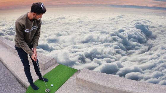 Man in a casual sweatshirt and dark pants stands on a small green practice mat on a rooftop, preparing a golf swing with clouds below at sunset.