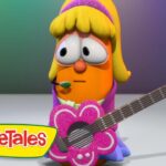 Orange VeggieTales character with blonde hair and a headset microphone, strumming a glittery pink flower-shaped guitar on a stage, VeggieTales logo in the corner.