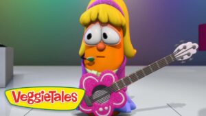 Orange VeggieTales character with blonde hair and a headset microphone, strumming a glittery pink flower-shaped guitar on a stage, VeggieTales logo in the corner.