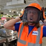 Person in blue shirt and orange safety vest lifts a face shield in a hardware store, with an excited expression.