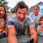 Man and four children smile as they lean in for a selfie around a round table in a backyard.
