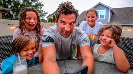 Man and four children smile as they lean in for a selfie around a round table in a backyard.