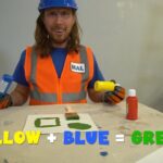 Man in a blue hard hat and orange safety vest demonstrates color mixing: yellow plus blue equals green, with painting supplies on the table.