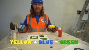 Man in a blue hard hat and orange safety vest demonstrates color mixing: yellow plus blue equals green, with painting supplies on the table.