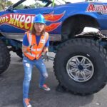 Person wearing an orange safety vest and blue hard hat gesturing in front of a large blue Hot Wheels monster truck with oversized tires.