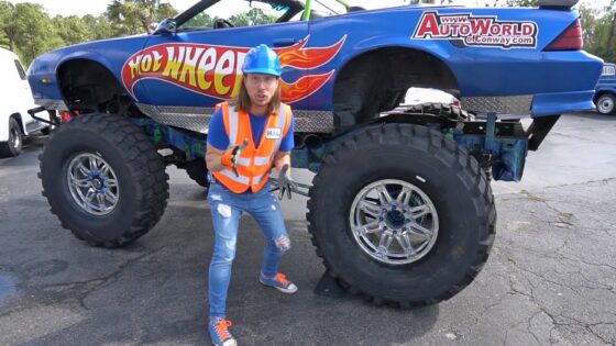 Person wearing an orange safety vest and blue hard hat gesturing in front of a large blue Hot Wheels monster truck with oversized tires.
