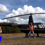 Person in orange safety vest and blue hard hat posing beside a black helicopter on a grassy airfield; cartoon worker illustration giving a thumbs up on the left.