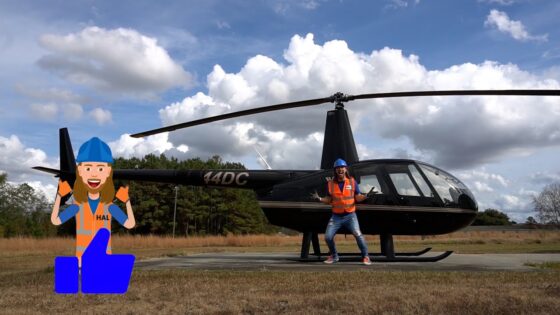 Person in orange safety vest and blue hard hat posing beside a black helicopter on a grassy airfield; cartoon worker illustration giving a thumbs up on the left.