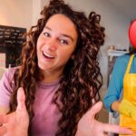 Young woman smiling and gesturing toward the camera in a bright cafe or classroom setting; a second person in a yellow apron stands behind with cleaning supplies and a red question-mark overlay on their face (mystery helper).