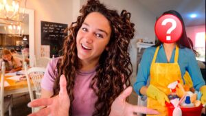 Young woman smiling and gesturing toward the camera in a bright cafe or classroom setting; a second person in a yellow apron stands behind with cleaning supplies and a red question-mark overlay on their face (mystery helper).