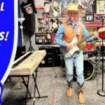 A man wearing a cowboy hat and vest plays a white electric guitar in a kids’ rock studio, with a large blue ‘School of Rock for Kids!’ banner and posters on the wall behind him (Cowboy Jack).