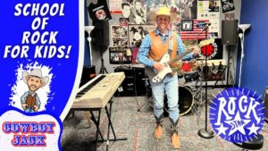 A man wearing a cowboy hat and vest plays a white electric guitar in a kids’ rock studio, with a large blue ‘School of Rock for Kids!’ banner and posters on the wall behind him (Cowboy Jack).