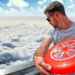 Man wearing sunglasses and a gray t-shirt holds a bright red frisbee with a white star logo on a railing above the clouds, looking to the side.