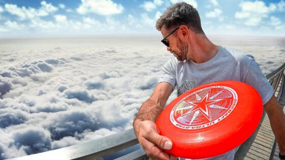 Man wearing sunglasses and a gray t-shirt holds a bright red frisbee with a white star logo on a railing above the clouds, looking to the side.