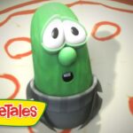 Larry the Cucumber from VeggieTales looks surprised, standing on a circular gray base with a red patterned background and the VeggieTales logo in the corner.