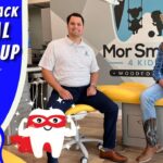 Two men sit on a yellow dental chair in a bright clinic, with a cowboy-themed banner reading 'Cowboy Jack Dental Check-Up' and a cartoon tooth character in front.