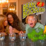 Group of kids at a kitchen table reacting to jelly bean tasting game, with a bright 'Bean Boozled' sign in the background.