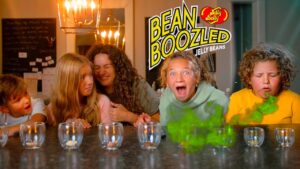 Group of kids at a kitchen table reacting to jelly bean tasting game, with a bright 'Bean Boozled' sign in the background.