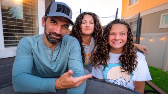 Three friends posing at a backyard table, smiling at the camera during sunset. The man on the left wears a blue shirt and cap; two girls sit beside him.