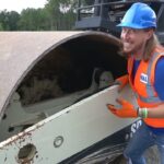 Construction worker in a blue hard hat and orange safety vest smiling beside a large road roller outdoors on a gravel lot.