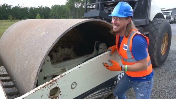 Construction worker in a blue hard hat and orange safety vest smiling beside a large road roller outdoors on a gravel lot.